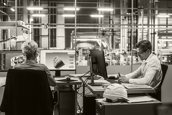 Two employees working at desks in front of computer monitors in an office inside a manufacturing plant. Source: Image via Getty Images