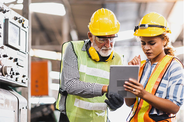 An expert and a novice are in an industrial setting, wearing yellow hard hats and safety vests. They are reviewing a workflow together on a tablet.